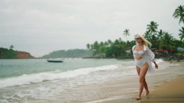 Slim blonde young woman in white shirt and onepiece swimsuit with straw hat wearing sunglasses walks along beach plays with hat. Happy pretty female walking by sea. Carefree girl enjoying summer