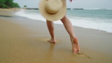 Slim female legs and feet walking along sea water waves on sandy beach. Pretty woman walks at seaside surf wearing white shirt carrying straw hat back view. Girl running in tide plays with wind