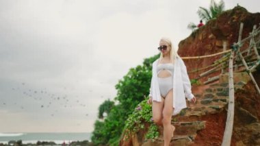 Young girl in swimsuit and white shirt walking barefoot down rocky hill. Blonde female walks down stairs cut in red rock. Young caucasian woman descending scenic hill using natural rocky stairs