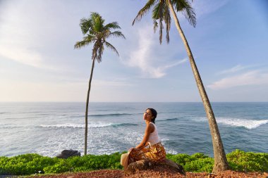 Young african female model posing in colorful clothes at scenic tropical location by ocean between palms at sunrise. Black woman sitting against exotic scenery with view of sea and palm trees at dawn
