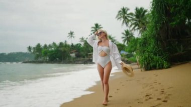 Slim blonde young woman in white shirt and onepiece swimsuit with straw hat wearing sunglasses walks along beach with palm trees on background gets splashed by wave. Happy pretty female walking by sea