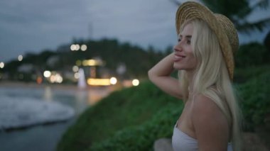 Young blonde woman in swimwear and straw hat on edge of picturesque hill with sea view and lights on background after sunset smiling enjoying the view alone. Caucasian young female in healing place