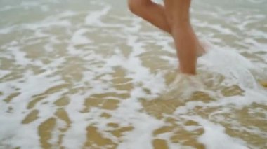 Slim female legs and feet walking along sea water waves on sandy beach. Pretty woman walks at seaside surf. Girl legs walking along shore on sand in tide leaving footprints carrying a straw hat