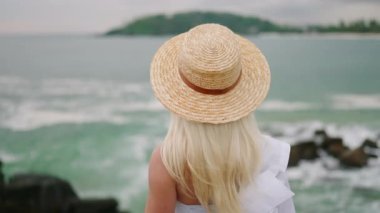 Young blonde woman sitting alone on bench self reflecting with spectacular view of ocean view from back. Blonde girl in white gown relaxes on bench at scenic tropical seaview location enjoying view