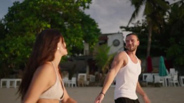 Young biracial happy couple walk holding hands and talking on the beach enjoying summer. Cheerful boyfriend and girlfriend relaxing and taking a walk at seaside hugging and kissing at sunrise