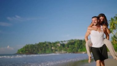 Boyfriend carrying girlfriend on his back, having fun at the seaside on summer day. Happy cheerful laughing couple on the beach fooling around and biggybacking. Young woman riding her mans back