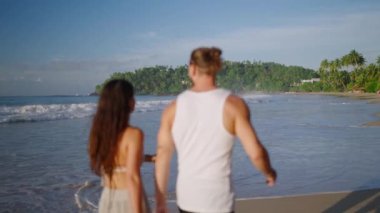 Young biracial happy couple unfocused walking on the beach together enjoying summer backview shot. Smiling boyfriend and girlfriend relaxing and taking walk at seaside at sunrise back shot.