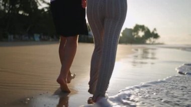 Legs of happy couple holding hands and walking on the beach together enjoying summer back view. Feet of heerful boyfriend and girlfriend relaxing and taking walk at the seaside at sunrise back shot.