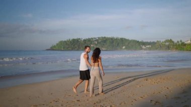 Young biracial happy couple walking on the beach together enjoying summer backview. Cheerful boyfriend and girlfriend relaxing and taking walk at seaside hugging and kissing at sunrise back shot