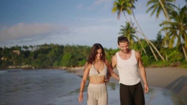 Young biracial happy couple holding hands and walking on the beach together enjoying summer. Cheerful boyfriend and girlfriend relaxing and taking walk at the seaside hugging and kissing at sunrise