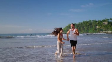 Young happy biracial couple holding hands running fooling around and dancing on the beach together enjoying summer back view. Biethnic boyfriend and girlfriend having fun at seaside at sunrise