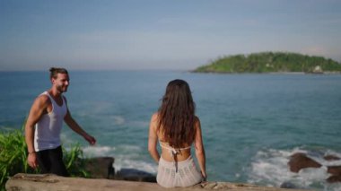 Guy approaches girl sitting alone on bench in front of sea and hugs cinematic shot. Man comes to bench by ocean with woman sitting, embraces her. Couple on bench at the edge of scenic hill back shot
