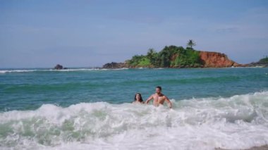 Young cheerful biracial couple in swimsuits holding hands running out of ocean waves splashing them slow motion shot. Multiracial happy man and woman run joyfully out of sea in slowmo front view