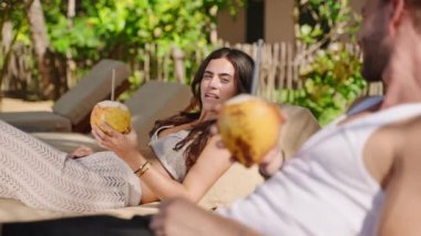 Young happy multiethnic couple relaxing on sunbeds at tropical resort clink and drink coconut water. Cheerful biracial tourists on summer vacation have two fresh king coconuts with exotic background
