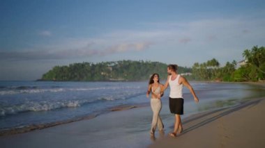 Young biracial happy couple holding hands and walking on the beach together enjoying summer. Cheerful boyfriend and girlfriend relaxing and taking walk at the seaside hugging and kissing at sunrise
