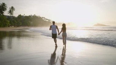 Silhouettes of young happy couple holding hands and running on the beach together enjoying summer back view. Boyfriend and girlfriend relax and walk at the seaside hugging and kissing at sunrise