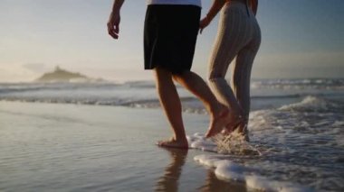 Legs of happy couple holding hands and walking on the beach together enjoying summer side view. Feet of heerful boyfriend and girlfriend relaxing and taking walk at the seaside at sunrise side shot.