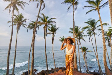 Young african female model posing in colorful clothes at tropical setting at sunrise. Black woman against exotic scenery at dawn. Multiracial dark-skinned model poses in front of palm trees at sunset