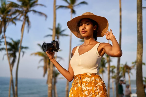Young african female model tourist with camera in colorful clothes and straw hat taking pictures at tropical location at sunrise. Black travelling woman takes photos in exotic ocean scenery at dawn