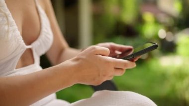 Unrecognizable woman using smart phone texting with AI, surfing social media, checking news, playing mobile games or texting messages sitting otside. Female spends time with cell gadget close-up
