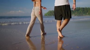 Legs of happy couple holding hands and walking on the beach together enjoying summer back view. Feet of heerful boyfriend and girlfriend relaxing and taking walk at the seaside at sunrise back shot.