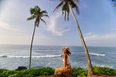 Young african female model posing in colorful clothes at scenic tropical location by ocean between palms at sunrise. Black woman sitting against exotic scenery with view of sea and palm trees at dawn