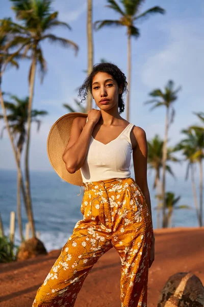 Young african female model posing in colorful clothes at tropical setting at sunrise. Black woman against exotic scenery at dawn. Multiracial dark-skinned model poses in front of palm trees at sunset