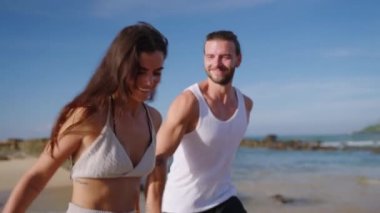 Young happy couple runs holding hands on beach through waves splashing water and enjoying summer closeup shot. Boyfriend and girlfriend having fun at spectacular scenic seaside tropical location.