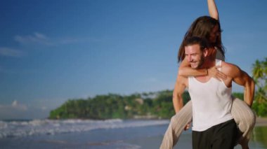 Boyfriend carrying girlfriend on his back, having fun at the seaside on summer day. Happy cheerful laughing couple on the beach fooling around and biggybacking. Young woman riding her mans back