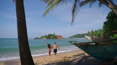 Young cheerful biracial couple in swimsuits holding hands running into the ocean splashing in waves slow motion backview shot. Multiracial happy man and woman run joyfully into sea in slowmo back shot