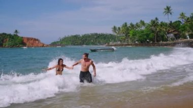 Young cheerful biracial couple in swimsuits holding hands running out of ocean waves splashing them slow motion shot. Multiracial happy man and woman run joyfully out of sea in slowmo front view
