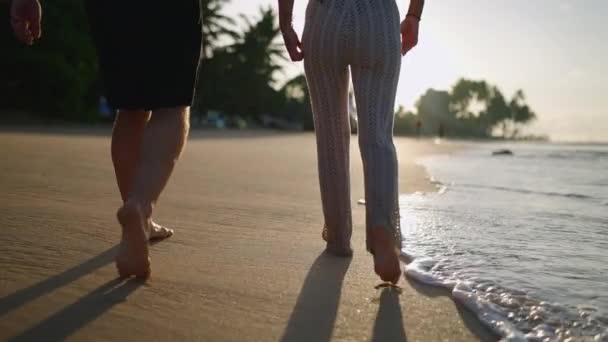 幸せなカップルの足が手を取り合い 夏のバックビューを楽しんで一緒にビーチを歩く 陽気なボーイフレンドとガールフレンドの足リラックスして取りますでザ海辺で日の出に戻るショット — ストック動画