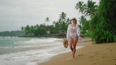 Slim blonde young woman in white shirt and onepiece swimsuit with straw hat wearing sunglasses walks along beach with palm trees on background. Happy pretty female walking by sea. Summer vacation