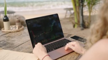 Woman cryptocurrency trader at laptop checking charts online working remotely at outdoor tropical seaside cafe. Female crypto broker analyses graphic of stock exchange rates and bets by ocean close-up