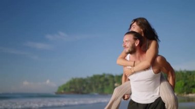 Boyfriend carrying girlfriend on his back, having fun at the seaside on summer day. Happy cheerful laughing couple on the beach fooling around and biggybacking. Young woman riding her mans back