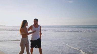 Young biracial happy couple holding hands and walking on beach together enjoying summer sideview. Cheerful boyfriend and girlfriend relaxing and talking at seaside hugging and kissing at sunrise.
