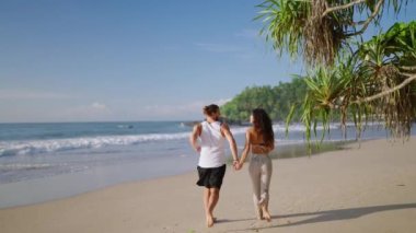 Young happy biracial couple holding hands running fooling around and dancing on the beach together enjoying summer back view. Biethnic boyfriend and girlfriend having fun at seaside at sunrise