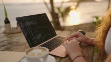 Dizüstü bilgisayardaki kadın kripto para tüccarı Candlestick çizelgelerini kontrol ediyor. Açık hava tropikal deniz kenarı kafesinde çalışıyor. Kadın kripto komisyoncusu hisse senedi kurlarını ve bahisleri okyanusa yakın hesaplıyor.