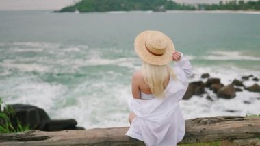 Young blonde woman sitting alone on bench self reflecting with spectacular view of ocean view from back. Blonde girl in white gown relaxes on bench at scenic tropical seaview location enjoying view