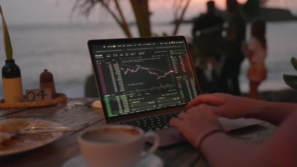 笔记本电脑上的妇女加密货币交易者在日落时在室外热带海滨咖啡馆远程工作 女性密码经纪人分析按海洋特写分列的证券汇率图表 — 图库视频影像