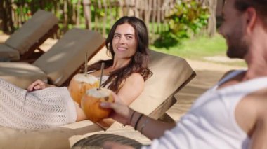Young happy multiethnic couple relaxing on sunbeds at tropical resort clink and drink coconut water. Cheerful biracial tourists on summer vacation have two fresh king coconuts with exotic background
