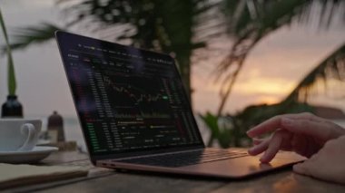 Dizüstü bilgisayardaki kadın kripto para tüccarı Candlestick çizelgelerini kontrol ediyor. Açık hava tropikal deniz kenarı kafesinde çalışıyor. Kadın kripto komisyoncusu hisse senedi kurlarını ve bahisleri okyanusa yakın hesaplıyor.