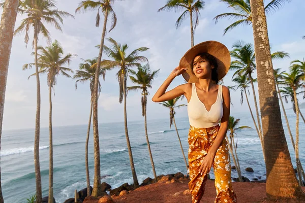 Young african female model posing in colorful clothes at tropical setting at sunrise. Black woman against exotic scenery at dawn. Multiracial dark-skinned model poses in front of palm trees at sunset