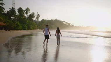 Genç mutlu çiftin siluetleri el ele tutuşup sahilde birlikte yürüyor ve yaz manzarasının tadını çıkarıyorlar. Erkek arkadaş ve kız arkadaş rahatlar ve gün doğumunda sahilde yürür sarılır ve öpüşürler.