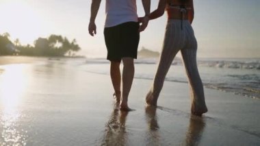 Legs of happy couple holding hands and walking on the beach together enjoying summer back view. Feet of heerful boyfriend and girlfriend relaxing and taking walk at the seaside at sunrise back shot.
