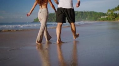 Legs of happy couple holding hands and walking on the beach together enjoying summer back view. Feet of heerful boyfriend and girlfriend relaxing and taking walk at the seaside at sunrise back shot.