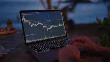 Dizüstü bilgisayardaki kadın kripto para tüccarı Candlestick çizelgelerini kontrol ediyor. Açık hava tropikal deniz kenarı kafesinde çalışıyor. Kadın kripto komisyoncusu hisse senedi kurlarını ve bahisleri okyanusa yakın hesaplıyor.