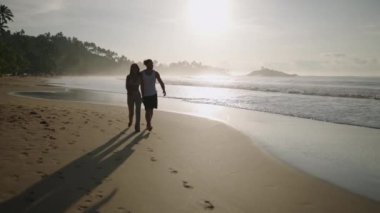 Çift ırklı mutlu genç çift el ele tutuşup sahilde birlikte yürüyor ve yaz manzarasının tadını çıkarıyor. Neşeli sevgili deniz kenarında dinleniyor ve konuşuyorlar. Gün doğumunda sarılıp öpüşüyorlar.