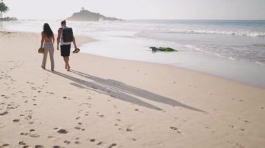 Young biracial happy tourist couple walking on the beach together enjoying summer backview shot from distance. Smiling boyfriend and girlfriend relaxing and taking walk at the seaside at sunrise.
