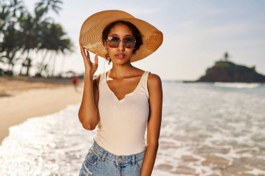 Hasır şapkalı ve güneş gözlüklü genç Afrikalı kadın model güneş doğarken tatil köyünde poz veriyor. Siyah kadın, kayalık yeşil adaya ve şafakta okyanus gelgitine karşı. Çok ırklı model tatilde poz veriyor